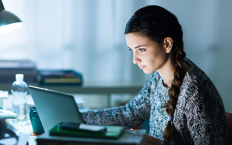 Woman sitting at a desk in a dimly-lit room, looking at a laptop screen.