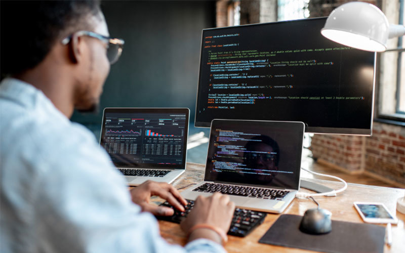 Young male business professional typing code. He is sitting at a desk with two laptops and one large monitor, all of which display code.