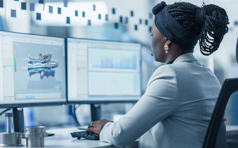 Young diverse female wearing a white blazer is working at a computer, designing a 3D model for the parts of some machine.