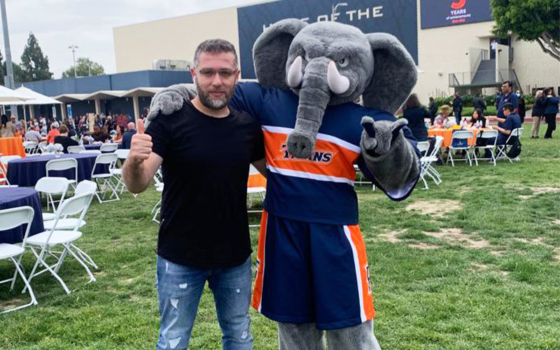 Muhittin Ceken posing for a picture with the Tuffy the Titan mascot on the Intramural Field at Cal State Fullerton. Muhittin is standing on the left giving a thumbs up, and Tuffy is to the right making a sign of the horns.
