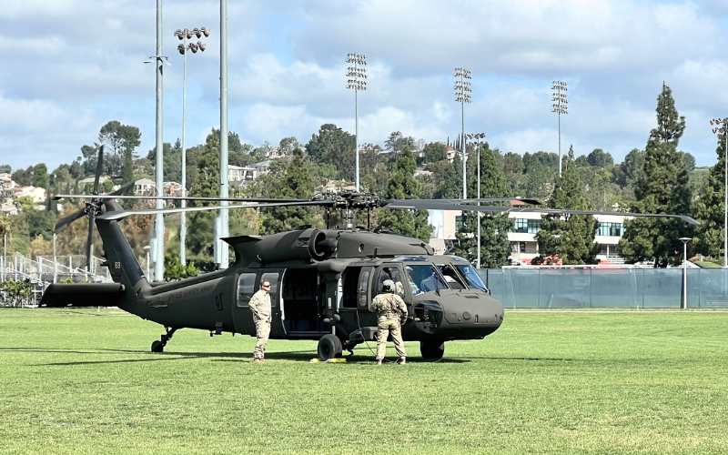 An Army H-60 helicopter sitting on the green grass of the CSUF Intramural Field with two Army personnel in uniform standing beside it.