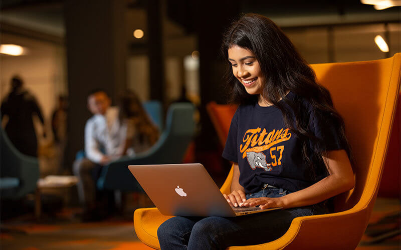 Female student wearing Titan gear typing on a laptop in Pollak Library.