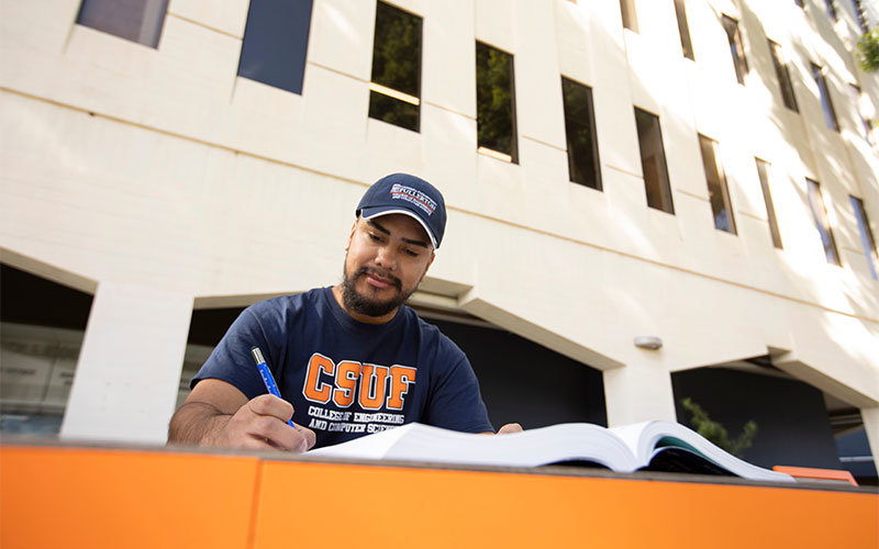 Student in Titan gear studying at an outdoor table next to the CSUF Engineering building.