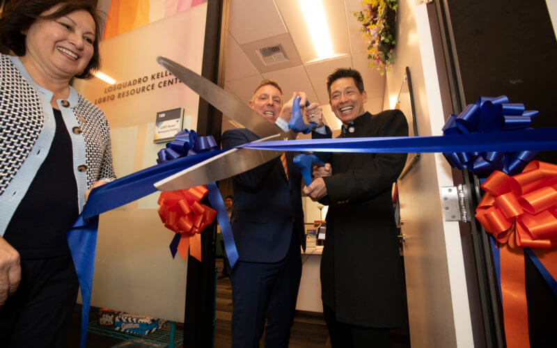 President Sylvia A. Alva watches Michael T. Losquadro and Dr. Brian C. Keller cut a blue and orange ribbon in the doorway of the Losquadro Keller LGBTQ Resource Center