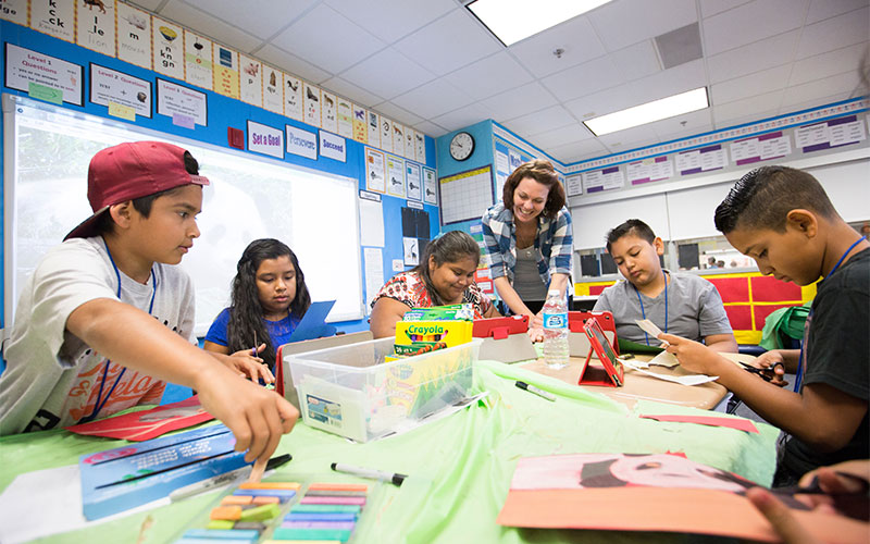 Female teacher in a classroom helping a group of five children work on arts and crafts projects.