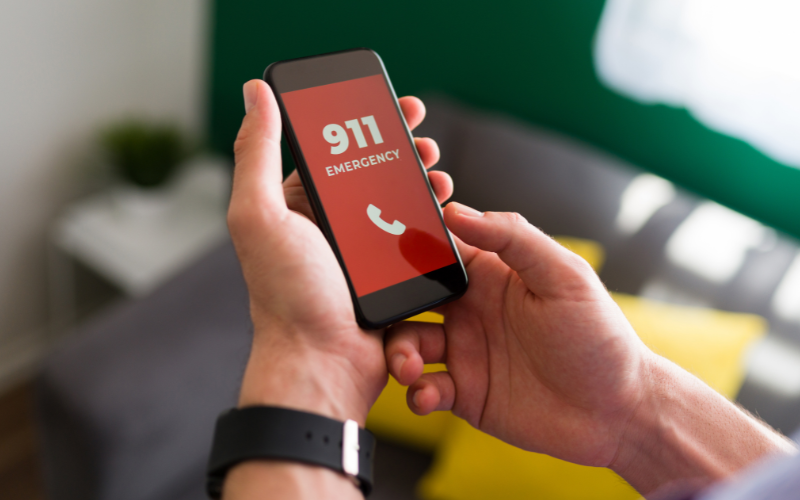 Person holding a cell phone in their hand. The screen is red with white lettering that says "911 Emergency."