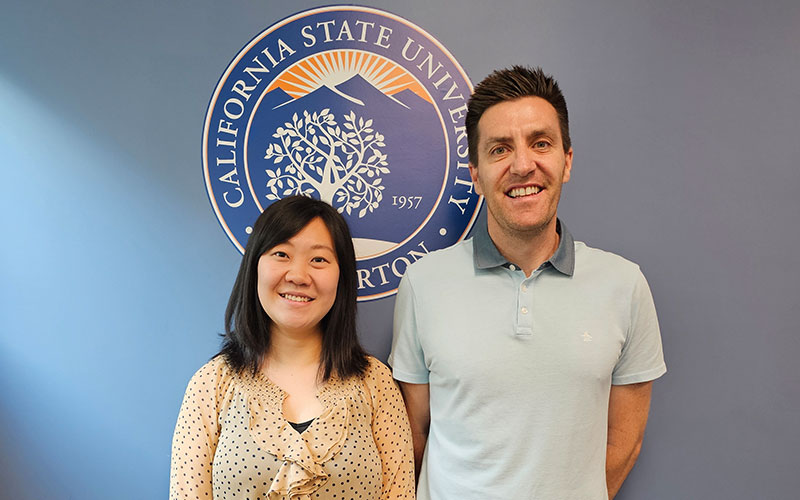 Dr. Yanan Feng and Dr. Matthew Llewellyn smiling in front of navy blue wall with large seal of Cal State Fullerton.