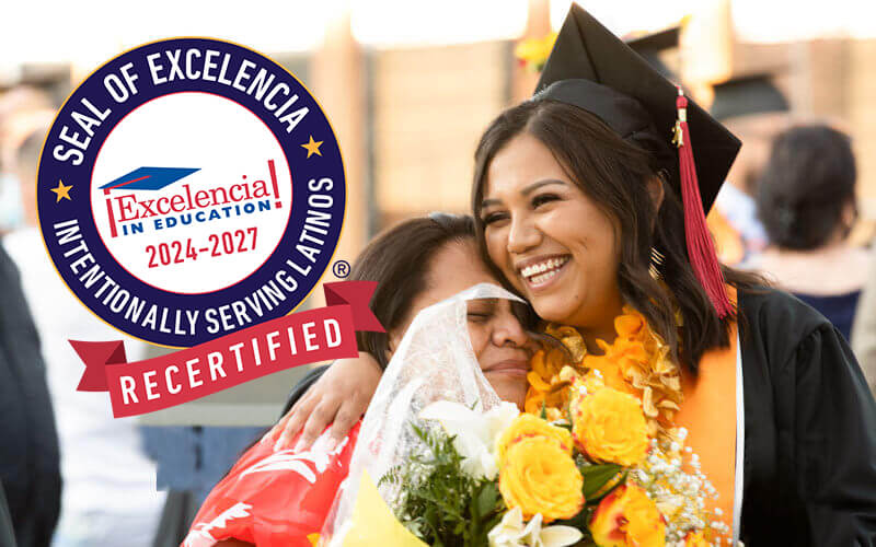 Graduate in cap and gown smiling and embracing a woman holding a bouquet of yellow flowers, alongside a circular logo reading ‘Seal of Excelencia, Intentionally Serving Latinos, Recertified 2024-2027’.