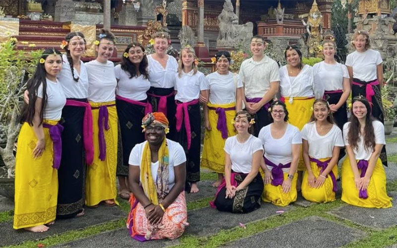 Group of diverse students wearing colorful skirts and sashes, posing in front of Buddhist temple in Thailand.