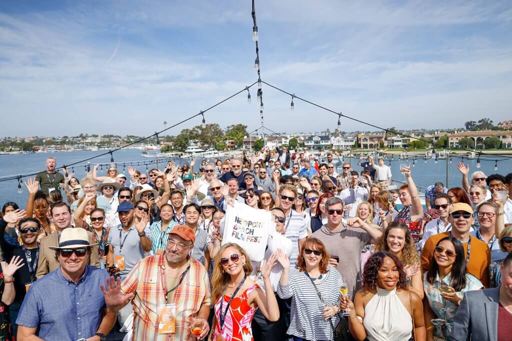 A large group of people smiling and waving at the camera during a sunny outdoor event by the water, with string lights overhead and houses in the background. Some attendees are holding drinks, and one person is holding a sign that reads ‘Newport Beach Film Fest.’