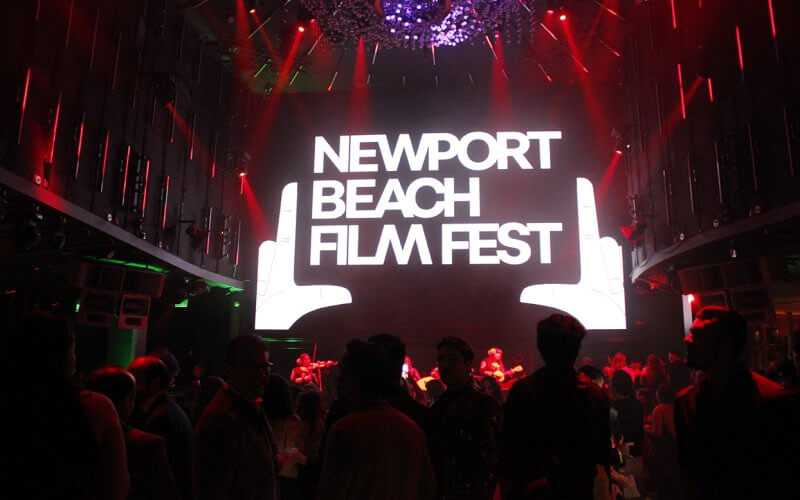 Audience in a dark room with a large illuminated screen reading 'Newport Beach Film Fest' and a hand gesture, surrounded by red lights and musicians performing on stage in the background.