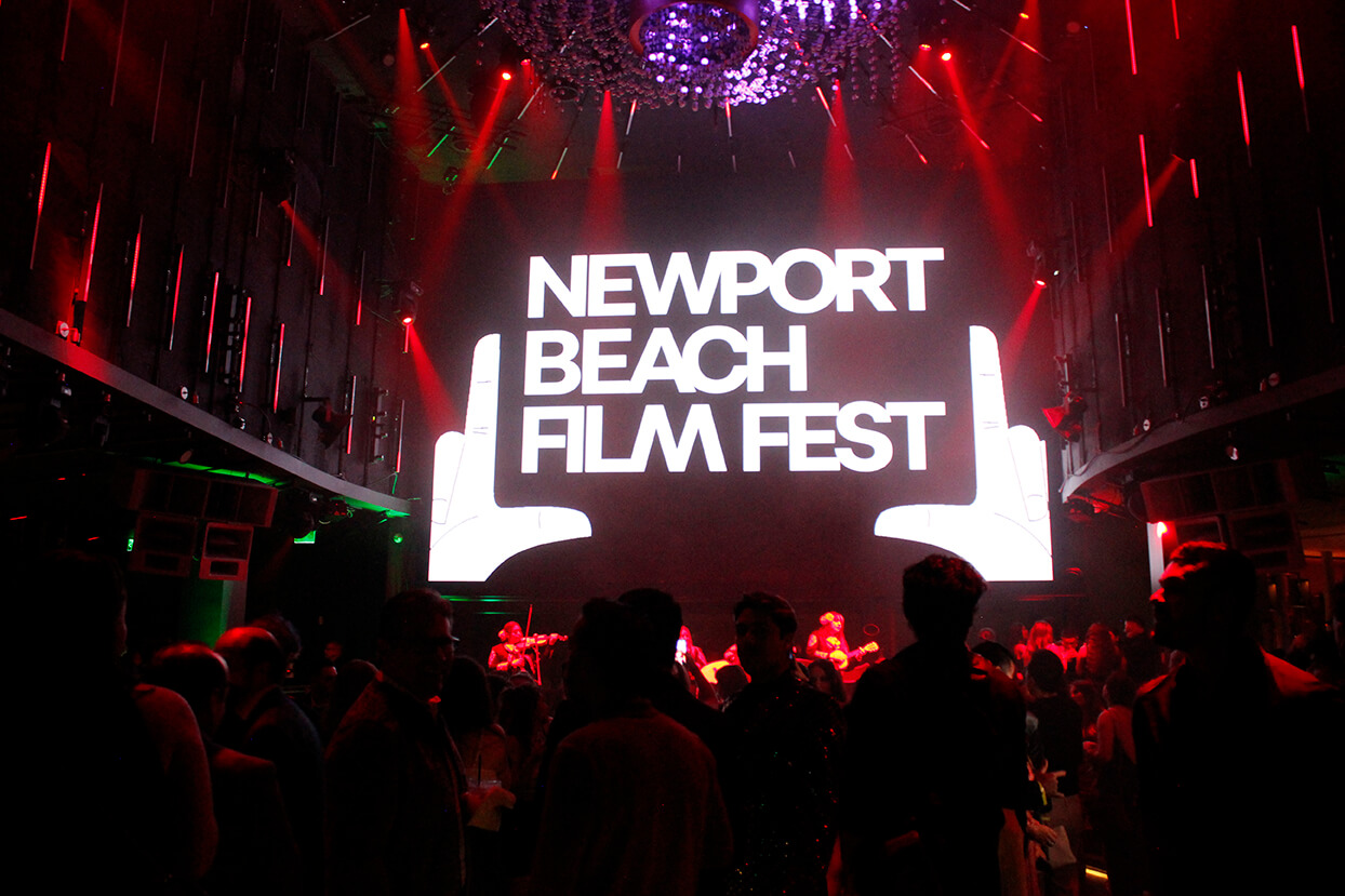 Audience in a dark venue with a large illuminated screen displaying ‘Newport Beach Film Fest’ and a hand gesture graphic, surrounded by red lights and musicians performing on stage in the background.