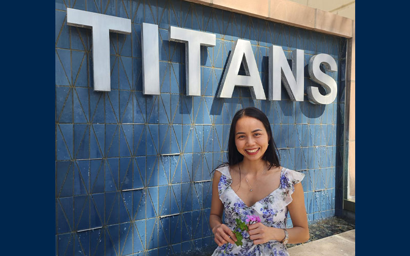 Image of Vui Nguyễn sitting in front of the TITANS fountain outside Pollak Library North. Vui is holding a small pink flower.