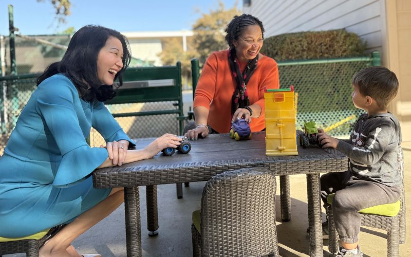 Two early childhood leaders smile and interact with a young boy as they play with toy cars.