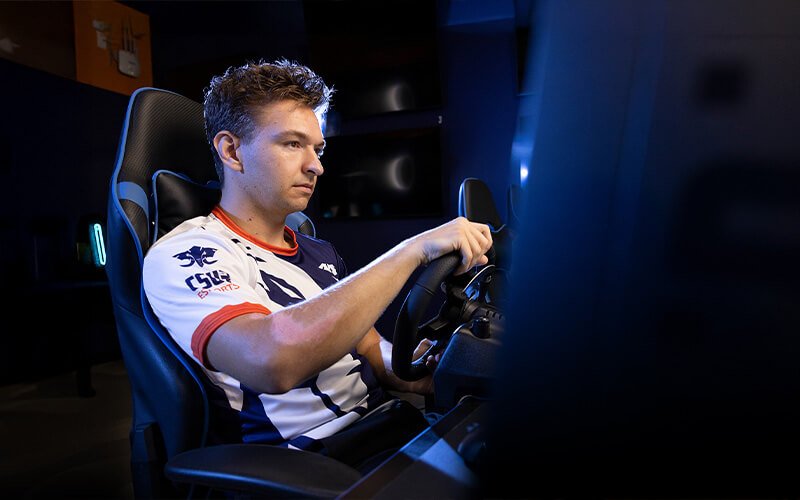 A man in a gaming jersey using a driving simulator setup with a steering wheel and monitor.