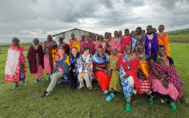 Lauren Heyden in a group photo featuring a diverse gathering of people, including individuals wearing vibrant, colorful traditional clothing. The group stands outdoors on green grass with a cloudy sky in the background.