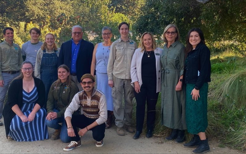 A group of 12 people smiling in front of a green background of trees