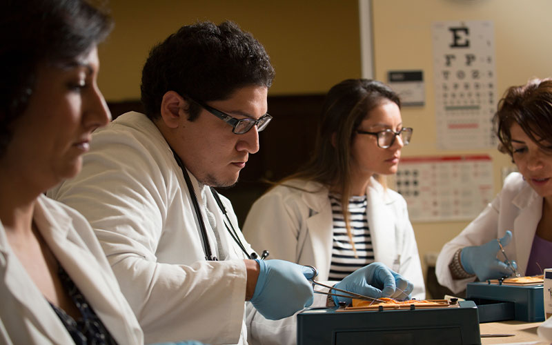 Four medical students in white lab coats practicing sutures