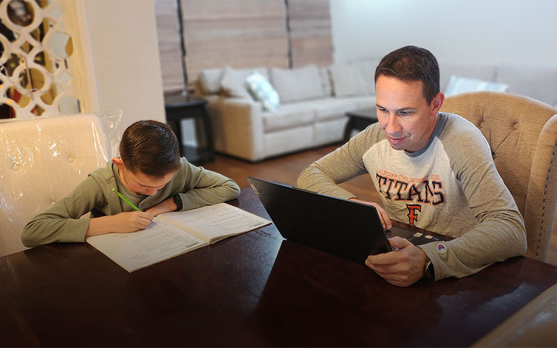 A young boy writes in a homework book while a man in a 'Titans' shirt uses a laptop beside him at a dining table.