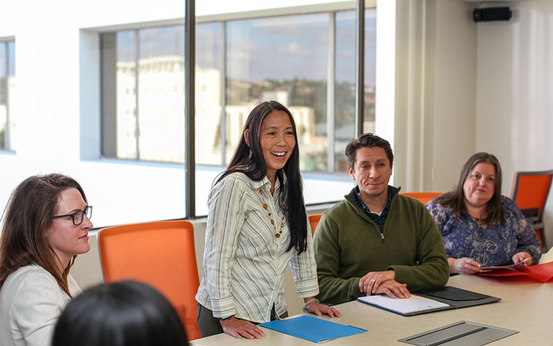 Group meeting in a conference room with large windows. Participants seated around a table with orange chairs.