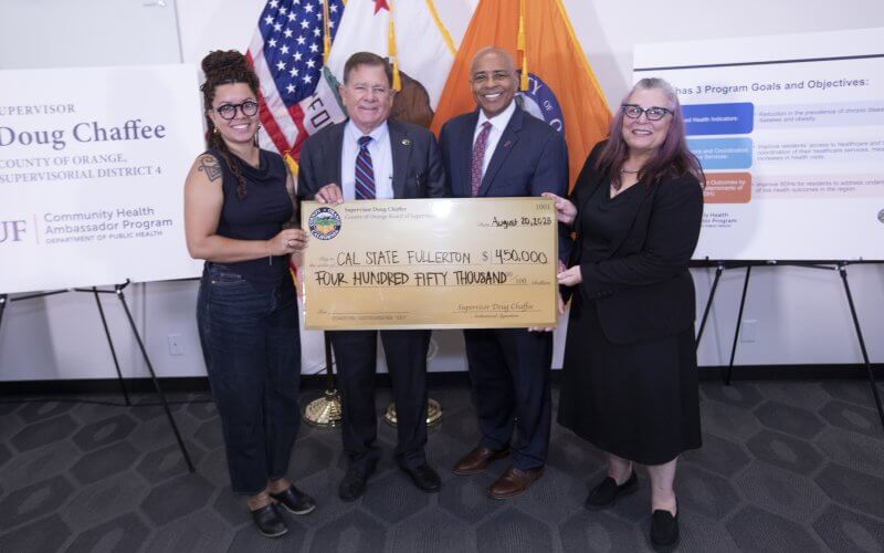 Cal State Fullerton officials and Orange County Board of Supervisors Chair Doug Chaffee pose with a giant check in front of County of Orange flags and signage