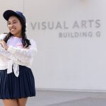 Skylar Williams standing outside the Visual Arts Building G, smiling with arms crossed, wearing a navy skirt, white textured blouse, and navy cap.