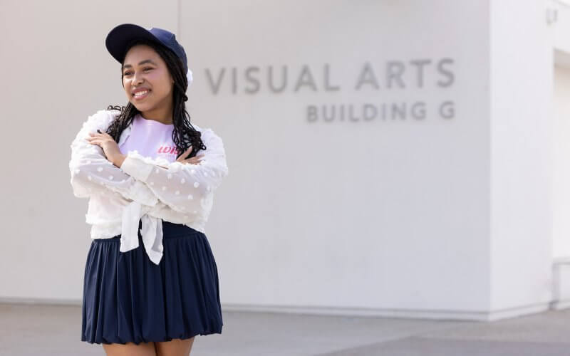 Skylar Williams standing outside the Visual Arts Building G, smiling with arms crossed, wearing a navy skirt, white textured blouse, and navy cap.
