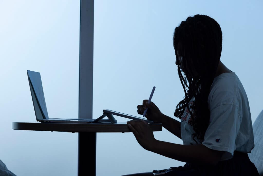 First-generation college student and aspiring storyboard artist Skylar Williams sitting at a table using a stylus on a tablet with a laptop open beside them, silhouetted against a bright window.