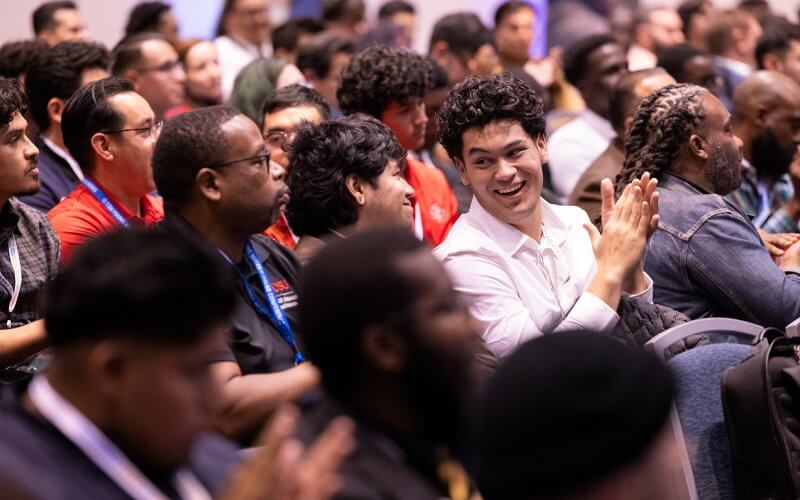 Audience members seated and engaged at the Young Males of Color Consortium Conference, with one attendee smiling and clapping.