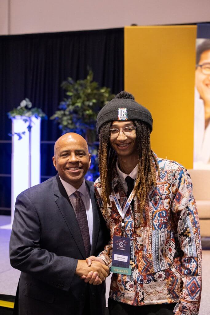 Two people smiling and shaking hands at the Young Males of Color Consortium Conference, one wearing a speaker badge.