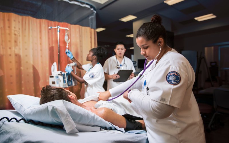 A nursing student checks a patient's chest with a stethoscope while two other nurses prepare an IV drip.