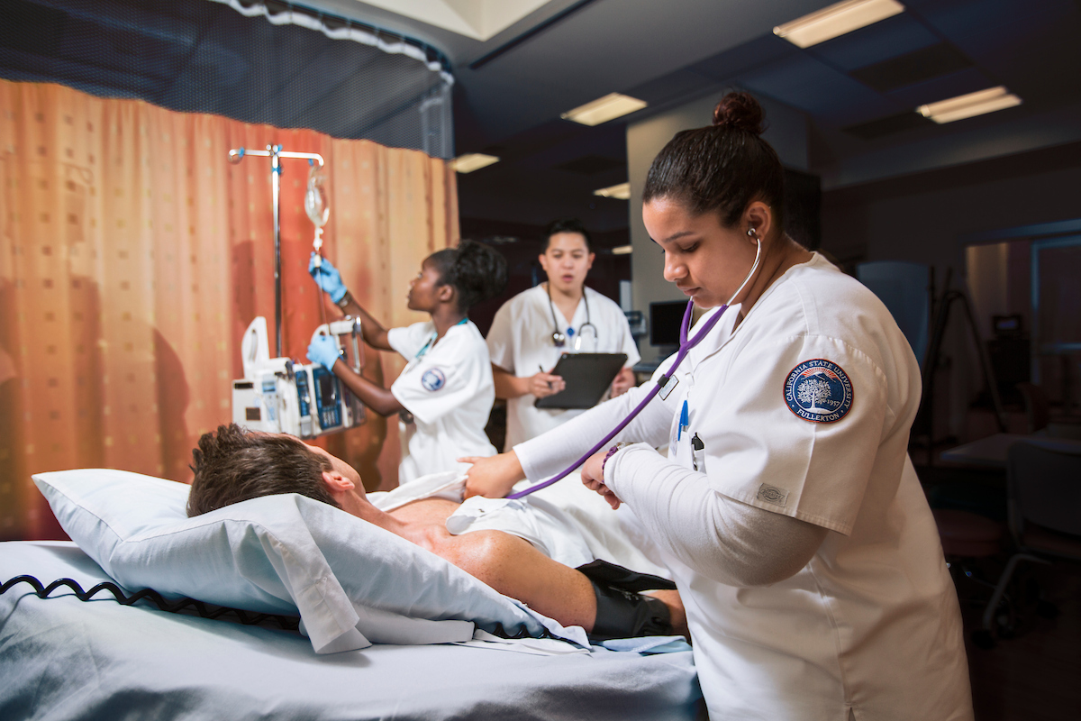A nursing student checks a patient's chest with a stethoscope while two other nurses prepare an IV drip.