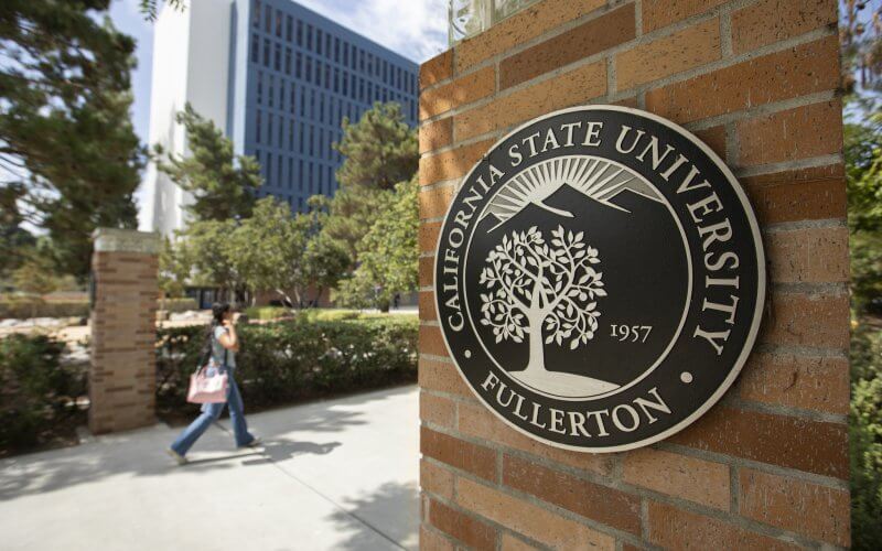 A student walks past a brick pillar displaying the California State University, Fullerton seal on a sunny campus walkway.