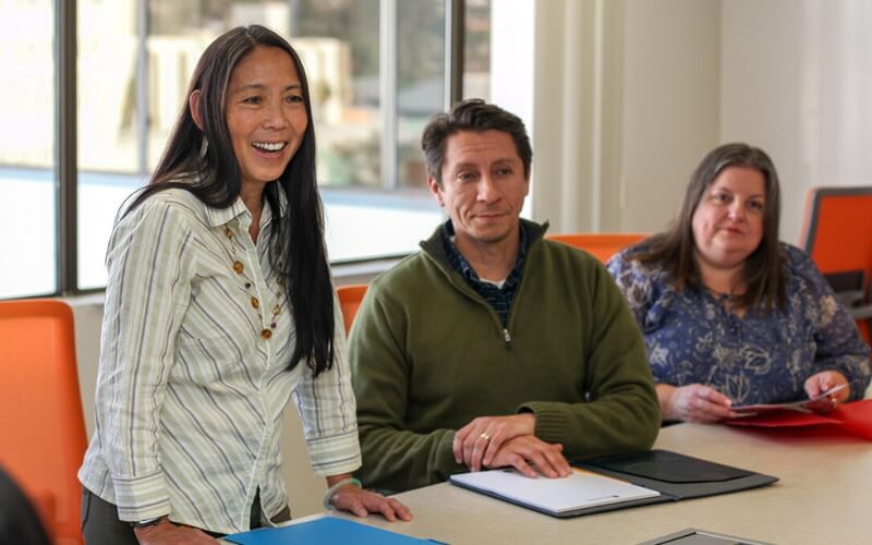Three people seated at a conference table in a meeting room, looking toward someone off camera; one person stands and smiles while speaking.