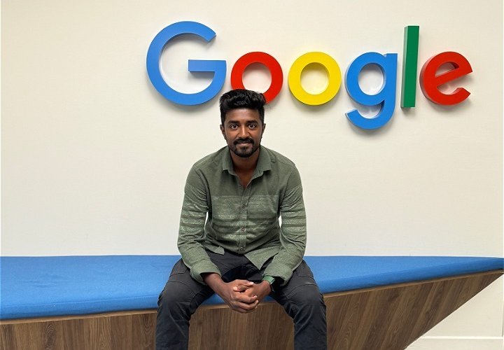 A CSUF graduate sits on a bench with the Google logo on the wall behind.