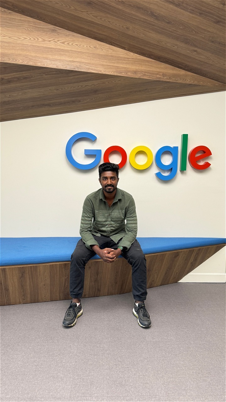 A CSUF graduate sits on a bench with the Google logo on the wall behind.