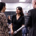 Three people standing in an office environment having a conversation; a woman in the center smiles while wearing a prosthetic arm as the others face her.