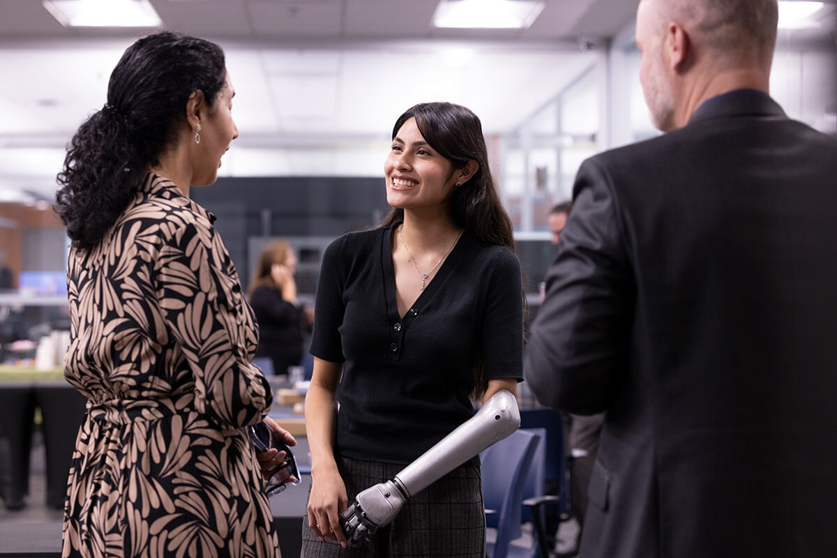 Three people standing in an office environment having a conversation; a woman in the center smiles while wearing a prosthetic arm as the others face her.
