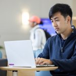 A student in a blue sweater sits at a table and works on a laptop in CSUF's College of Business and Economics