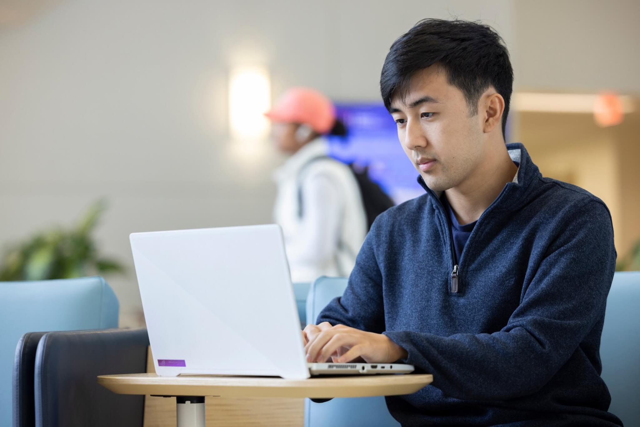 A student in a blue sweater sits at a table and works on a laptop in CSUF's College of Business and Economics