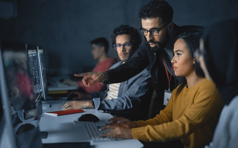 A person points over someone's shoulder at a computer.