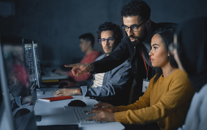 A person points over someone's shoulder at a computer.