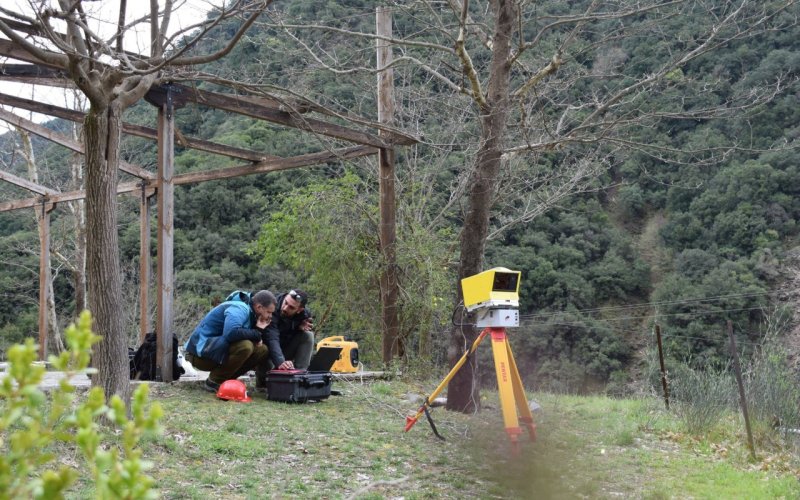 Two individuals are crouched on the grass, working with equipment in an outdoor setting with trees and hills. A yellow tripod with a device is positioned nearby.