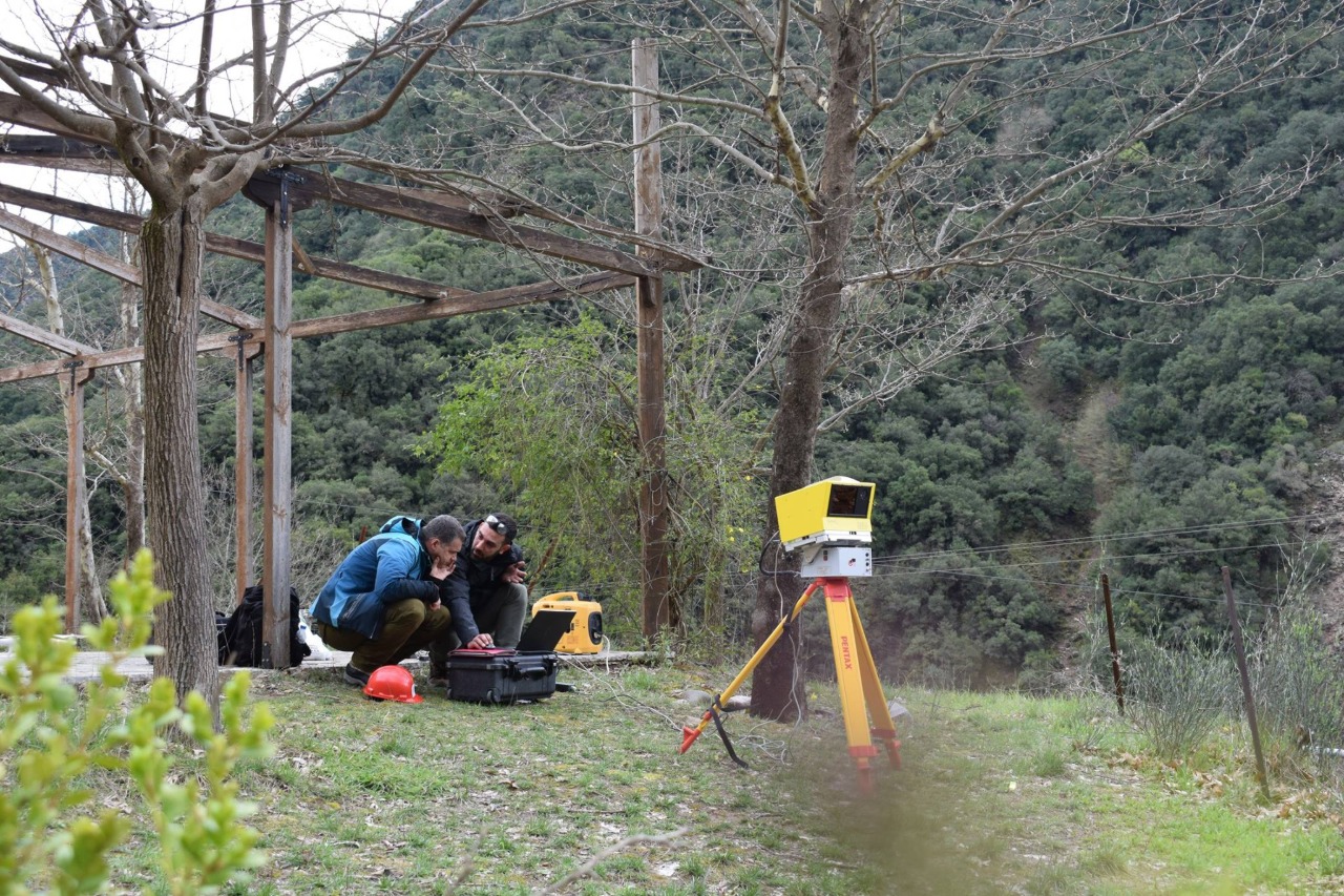Two individuals are crouched on the grass, working with equipment in an outdoor setting with trees and hills. A yellow tripod with a device is positioned nearby.