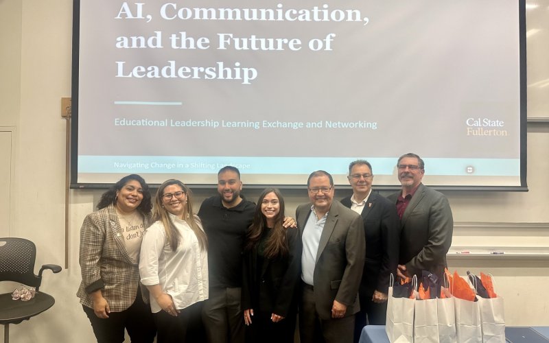 Seven panelists pose for a group photo in front of a presentation screen that reads, "AI, Communication, and the Future of Leadership"