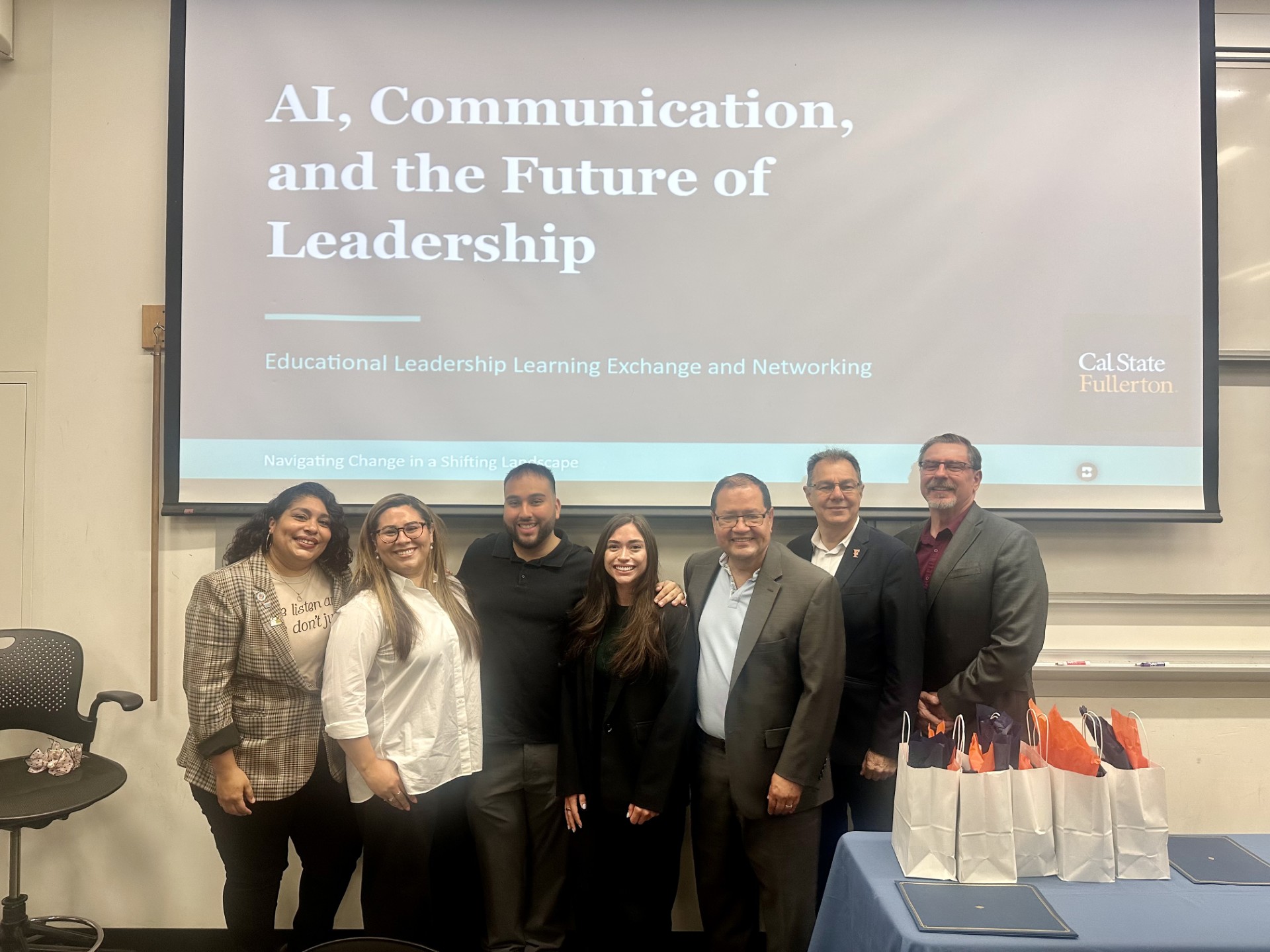 Seven panelists pose for a group photo in front of a presentation screen that reads, "AI, Communication, and the Future of Leadership"