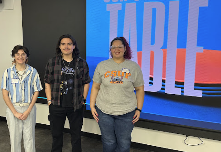 Three students stand in front of a presentation screen that reads, "Our Collective Table."