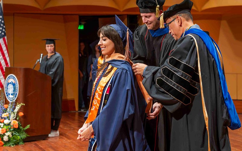A business graduate student is hooded by two faculty members at the Graduate Programs Hooding Ceremony.