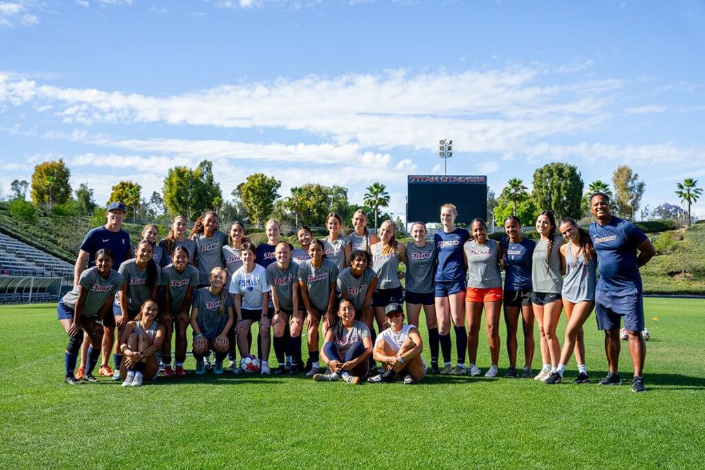 Group photo of a Cal State Fullerton women’s soccer team and coaches standing together on Titan Stadium field under a bright blue sky to spread the message of e-vehicle safety