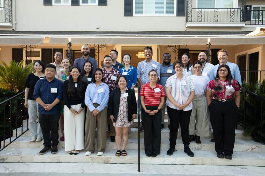 Group of newly hired faculty members pose together on the front steps of a campus residence during a welcome gathering, with the building entrance behind them.